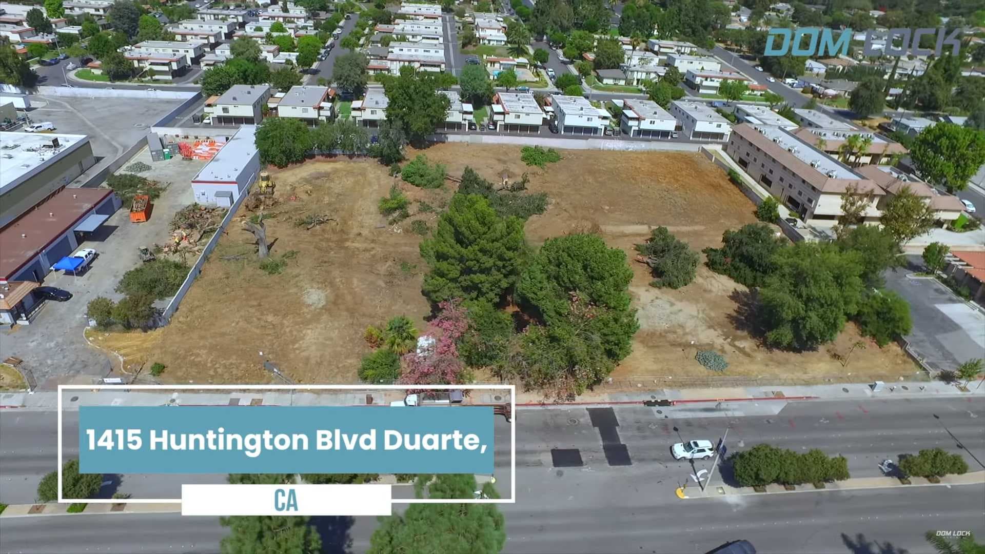 Aerial view of a block of houses and a dirt lot with trees; Dom Lock Aerials watermark