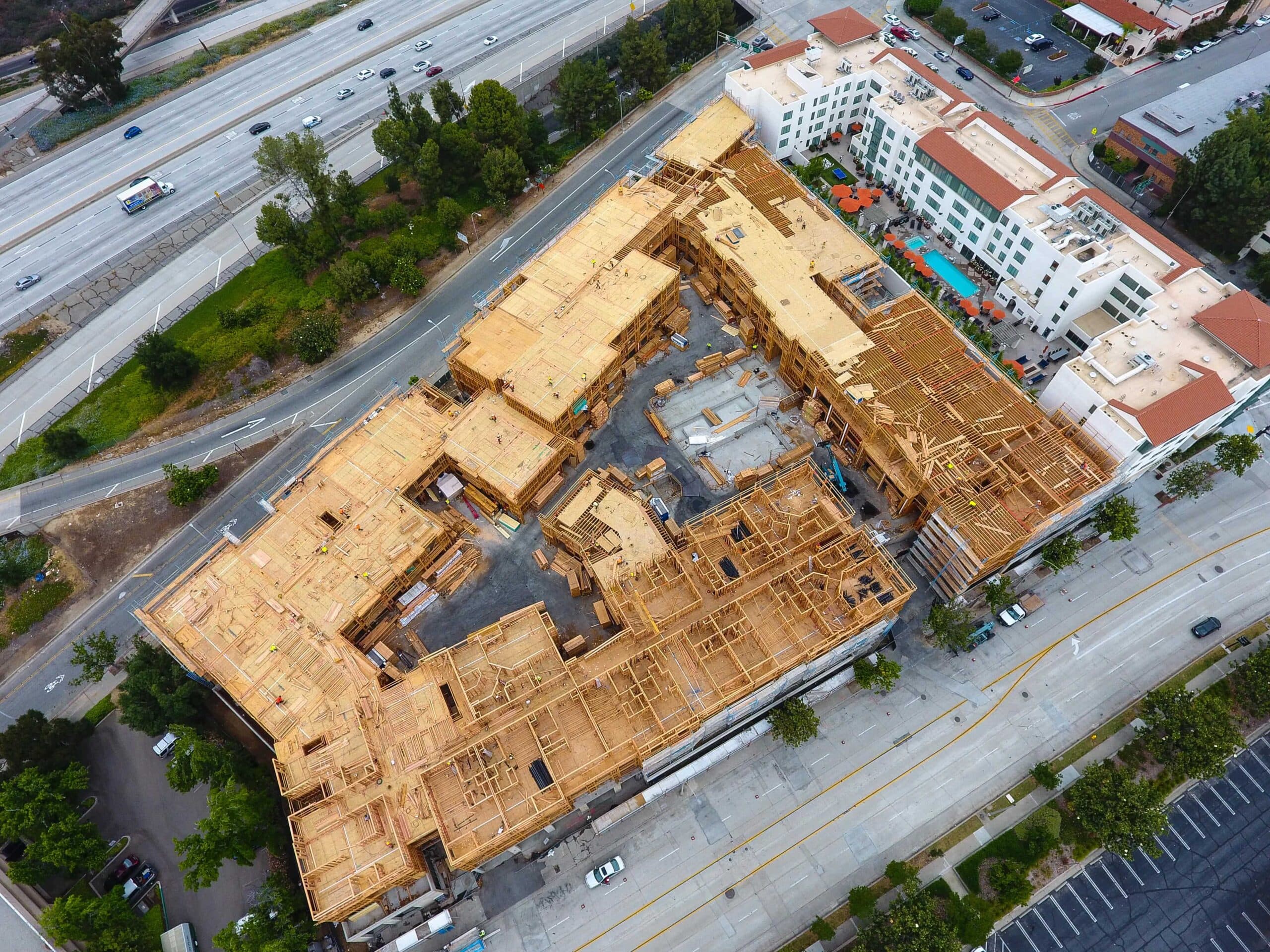 Aerial view of a wooden-framed construction site beside a highway, with trees and white modern buildings