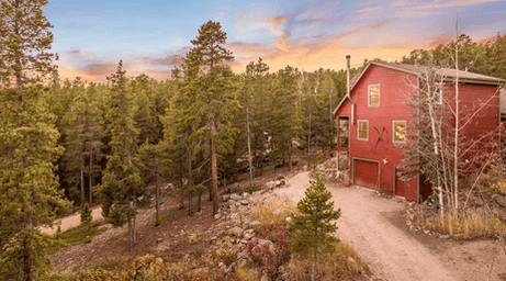 Red two-story house with a garage beside a dirt road, surrounded by tall pine trees at sunset