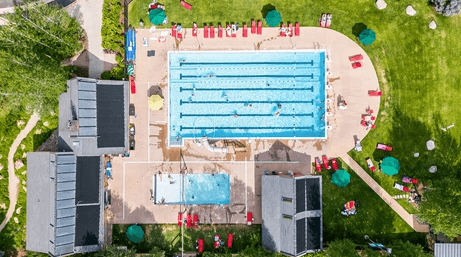 Aerial view of an outdoor pool complex with lanes, deck chairs, people around, and surrounding buildings