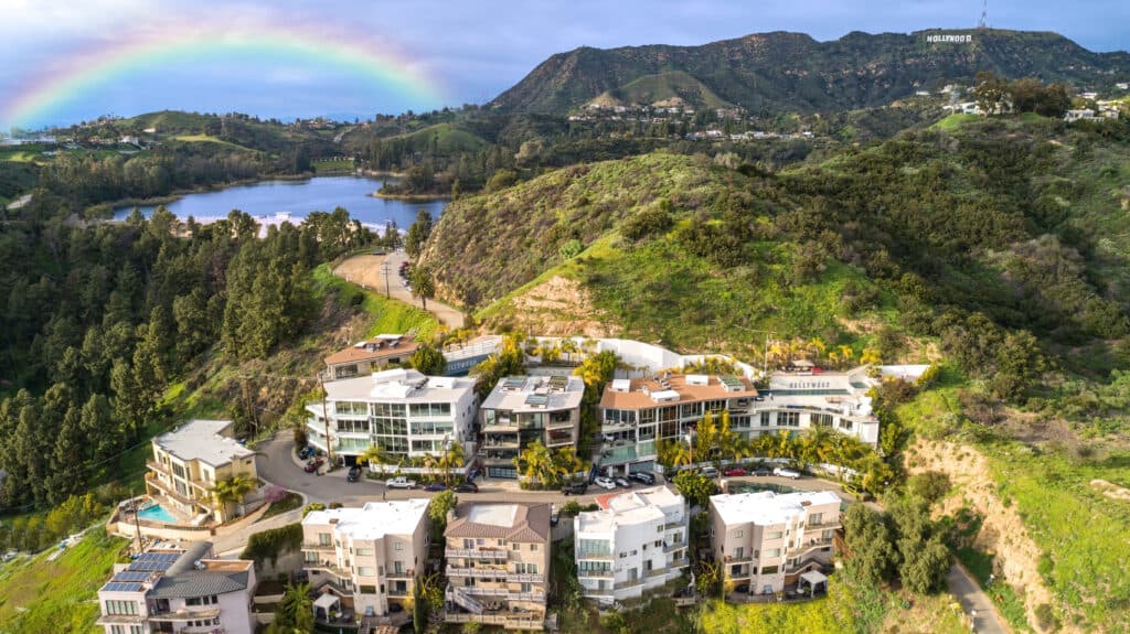 Hillside residential buildings clustered along a winding road, with trees, lake, rainbow, and Hollywood sign