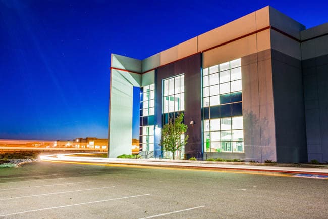 Modern building with glass windows and a blue-green feature, light trails across a parking lot at night