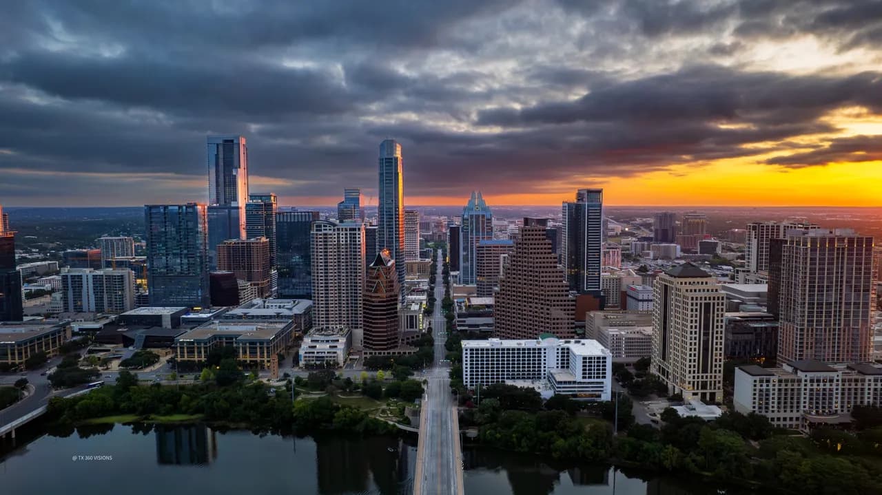 Downtown skyline at sunset with a river, orange sky, a bridge, and tall glass buildings