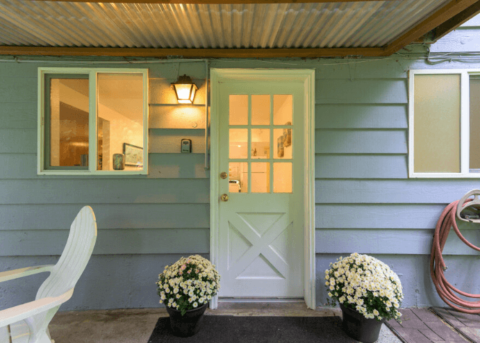 Blue front porch with white paneled door, two windows, wall lantern, a chair, and two pots of white mums