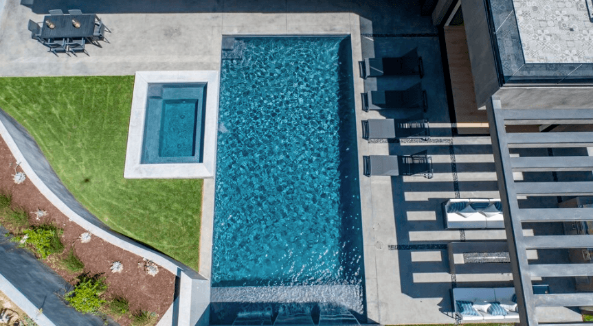 Aerial view of a modern backyard with a rectangular pool, a square spa, a green lawn, and outdoor seating