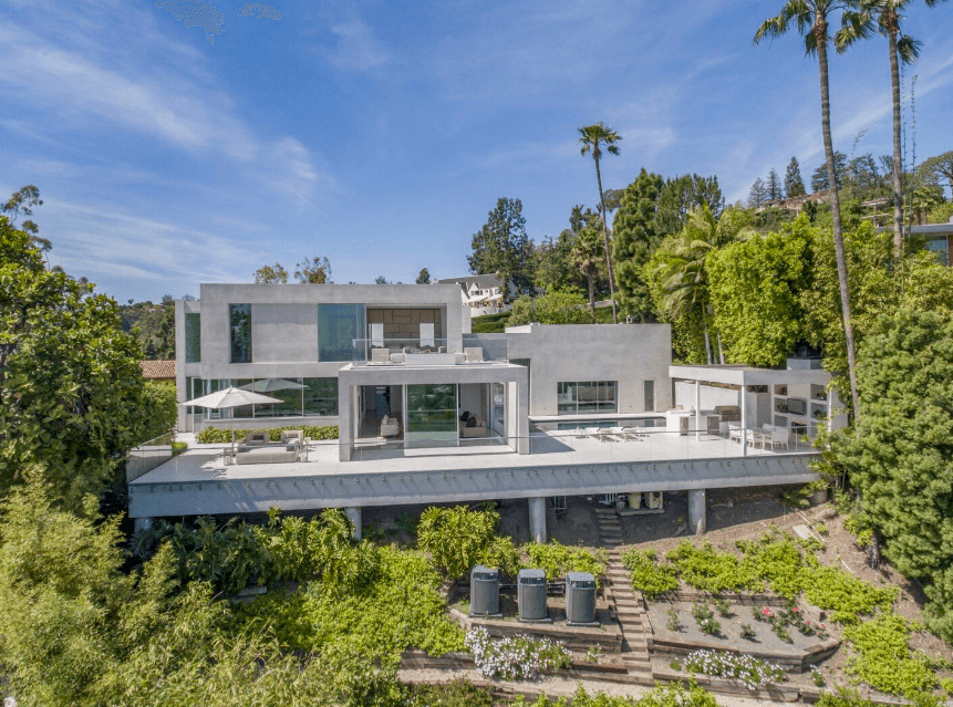 Modern two-story house with glass walls and a large deck, surrounded by palm trees and lush landscaping