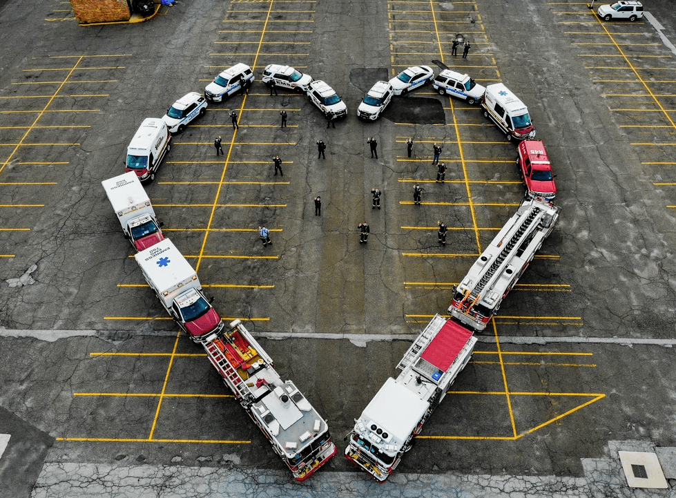 Aerial view of ambulances and fire trucks arranged in a heart shape in a parking lot with responders